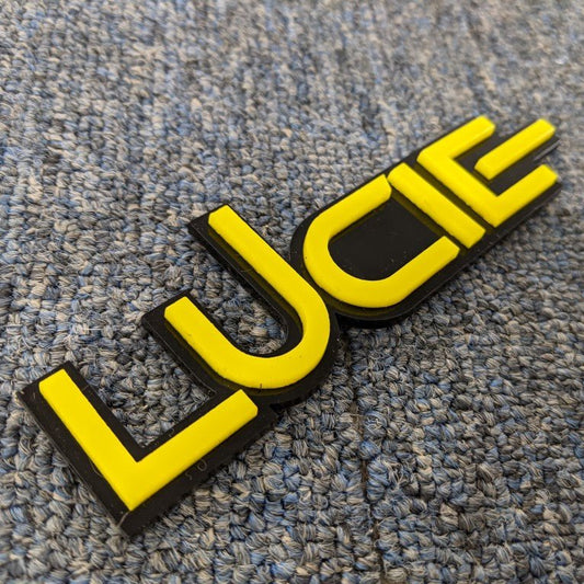 Lucie Car Badge - Yellow On Gloss Black - Squid Font - Tape Mount - Atomic Car Concepts