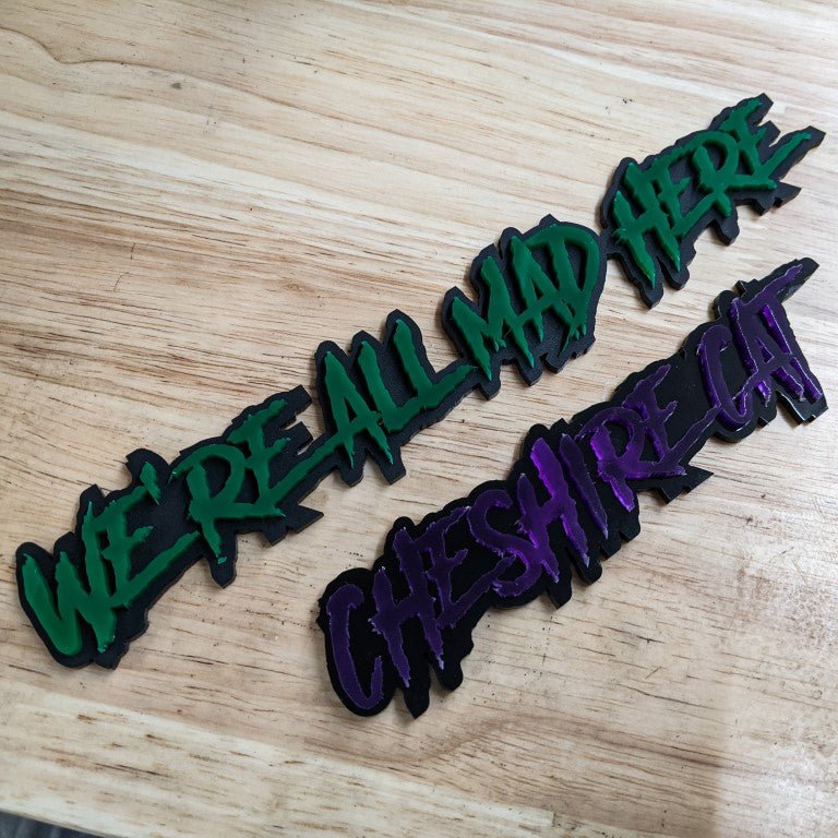 We're All Mad Here and Cheshire Cat Car Badge - Green And Purple On Matte Black - Aggressive Font - Tape Mounting - Atomic Car Concepts